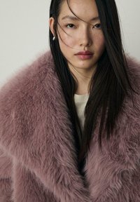 Puhast, svetlo roza umetno krzno plašč z širokim ovratnikom, ki ima mehak zunanji sloj in plastno zasnovo. Obrabljen čez kremasto pulover.