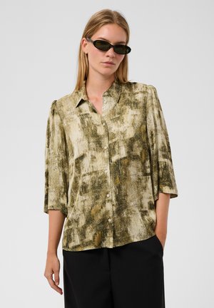 Mujer que lleva una blusa de botones con estampado en verde oliva, mangas hasta el codo, pantalones negros y pequeñas gafas de sol negras sobre un fondo liso.