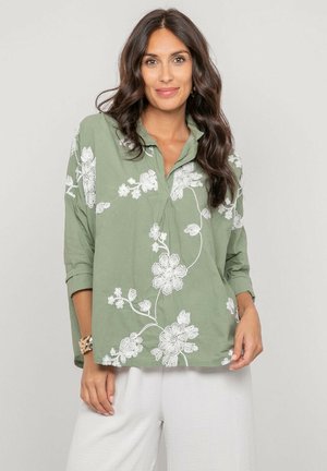 Femme aux longs cheveux foncés portant une chemise verte ample avec une broderie florale blanche et un pantalon blanc, debout devant un fond uni.