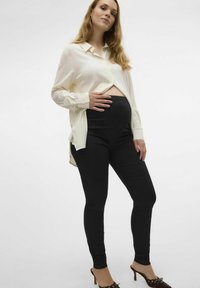 Hvit skjorte med knapper, lagd over svarte gravidleggings, som fremhever en jevn tekstur. Modellen poserer med en hånd på magen.