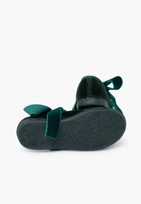 Zapatos de terciopelo verde con una suela negra texturada, con detalle de lazo y correas en el tobillo. La parte inferior presenta patrones florales.