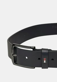 Cintura in pelle nera con fibbia in metallo nero lucido, cinturino regolabile con fori e un piccolo logo con accenti rossi e bianchi.