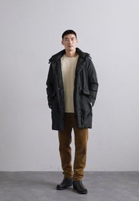 Caban negru cu puf și glugă, având un fermoar frontal, buzunare laterale și o textură matlasată. Purtați peste un pulover tricotat crem și pantaloni din catifea.