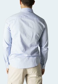 Camicia azzurra chiaro con maniche lunghe, texture morbida, orlo curvo e polsini con un solo bottone, indossata con pantaloni cargo beige.
