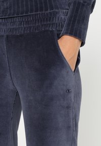 Navyblaue Velour-Sweatpants mit einem gerippten Bund, seitlichen Taschen und einem kleinen Logo-Akzent am Unterschenkel. Die weiche Textur ist sichtbar.