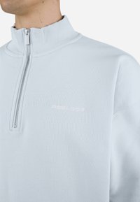 Lyseblå pullover med en kvart-lynlås krave, ribbet tekstur og subtilt logo i hvid på brystet. Blødt stof og afslappet pasform.
