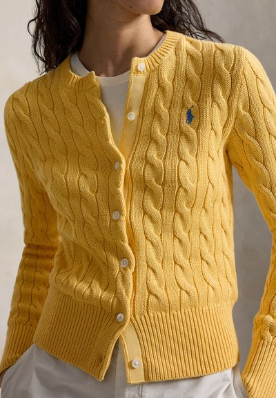 Femeie purtând un cardigan galben cu model împletit peste o cămașă albă, cu nasturi albi și un mic logo brodat albastru pe piept.