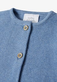 Strickjacke in Hellblau mit rundem Halsausschnitt, strukturiertem Stoff und zwei auffälligen Holzknöpfen an der Vorderseite. Markenlabel sichtbar.