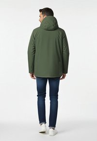 Oliivinvihreä vedenpitävä takki, jossa on huppu, yksinkertaisella muotoilulla ja kahdella sivutaskulla, yhdistetty tummansinisille farkuille ja valkoisille tennareille.