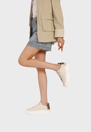 Donna con giacca beige, gonna di denim e sneakers bianche con suola platform e tacco leopardato, in piedi su una gamba su sfondo bianco.