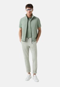 Hellgrüner ärmelloser Pullover über einem hellgrünen Polo, kombiniert mit hellgrauen Hosen und weißen Sneakers. Der Pullover hat einen hohen Kragen und einen Reißverschluss.