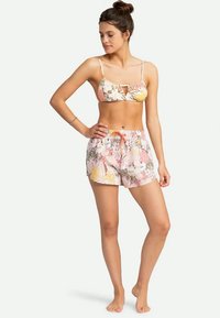 Žena stojící naboso, oblečená v bikinovém topu a odpovídajících šortkách s květinovým vzorem, s jednou rukou na bok a s pohledem skloněným na stranu.
