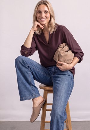 Femme aux cheveux blonds assise sur un tabouret en bois, portant un chemisier violet, un jean bleu, des chaussures marron sans lacets, et tenant un sac beige sous un bras.