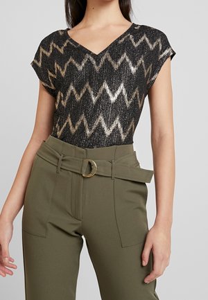 Schwarz und goldenes Zickzack-Muster-Oberteil mit V-Ausschnitt, kombiniert mit hoch taillierten olivgrünen Hosen mit einem Gürtel und metalischen Ringdetails.
