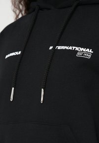 Sweatshirt preta com capuz e cordão. Apresenta texto branco da marca "Barbour International EST 1936." Textura de tecido lisa. Detalhes metálicos prateados.