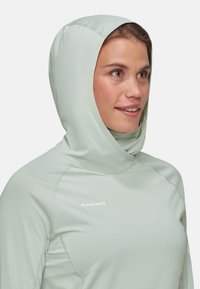 Ljust grön huva med slät textur, som har platta sömmar och en subtil logotyp på bröstet. Den figurnära designen ökar komforten.