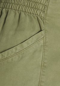 Zbliżenie na oliwkowo-zielony materiał z widocznym przeszytym elastycznym paskiem w talii oraz detalem bocznej kieszeni na odzieży.