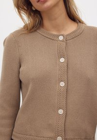 Cardigan en tricot beige avec un col rond, présentant un motif en chevron et cinq grands boutons blancs sur le devant.