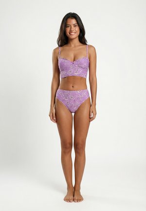 Jonge vrouw staat blootsvoets en draagt een lila bloemen tweedelige lingerie met geschulpte randen, glimlachend tegen een effen lichte achtergrond.