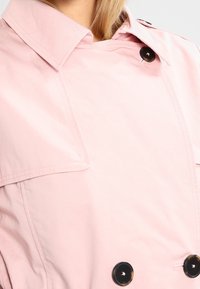 Jasnoróżowy płaszcz trencz z kołnierzem w szpic, dwurzędowym zapięciem na duże czarne guziki oraz pagonami na ramionach. Gładka faktura.