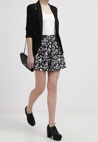 Blazer noir porté sur un haut blanc, associé à une jupe florale noir et blanc. Des chaussures noires à talons épais complètent le look.