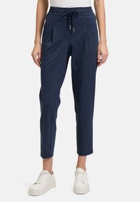 Marineblauwe joggers gemaakt van zachte, rekbare stof met een trekkoord in de taille, geplooide voorkant en taps toelopende benen. Gedragen met witte sneakers.