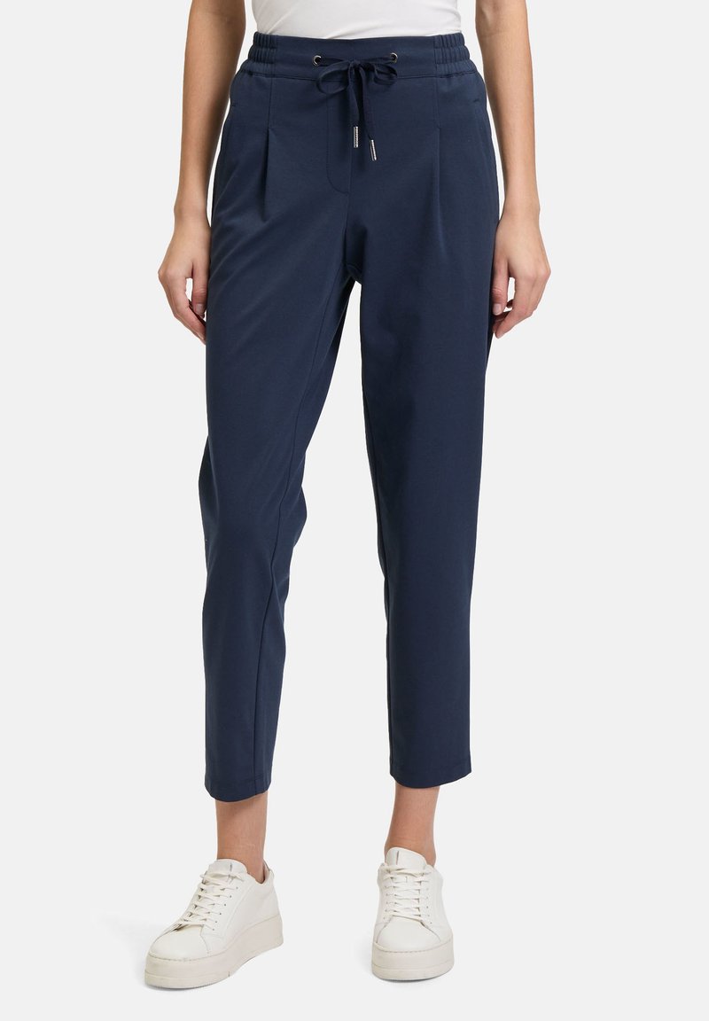 Marineblauwe joggers gemaakt van zachte, rekbare stof met een trekkoord in de taille, geplooide voorkant en taps toelopende benen. Gedragen met witte sneakers.