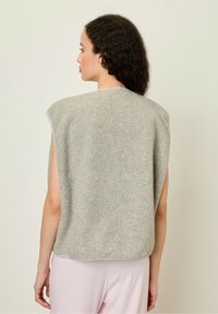 Světle šedý bezrukávový fleece top s texturovaným povrchem, kulatým výstřihem a surovými okraji. Nosený přes pastelově růžové spodní části.