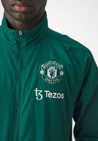 Chubasquero verde con cuello alzado, con el logo del Manchester United y "Tezos" impreso en blanco. El material parece ligero y tiene una textura suave.