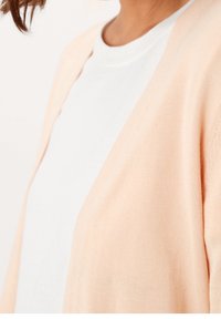 Brzoskwiniowy kardigan wykonany z miękkiego, lekkiego dzianinowego materiału o luźnym kroju, z dekoltem w serek i otwartym przodem.