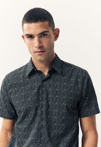 Košulja s kratkim rukavima tamnoplavih nijansi sa složenim svijetlim cvjetnim uzorkom. Ima špicasti ovratnik i zatvaranje na gumbe, izrađena od lagane tkanine.