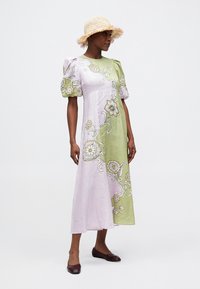 Vestido com mangas bufantes, design assimétrico em rosa e verde, com bordados florais intrincados, feito de tecido leve. Combinado com um chapéu de palha.