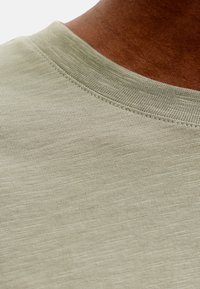 Κοντινό πλάνο στον λαιμό ενός T-shirt από υφασμάτινο υλικό σε ανοιχτό μπεζ χρώμα, σε άτομο με σκούρο χρώμα δέρματος.