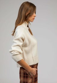 Pull en tricot couleur crème avec un col montant, des poignets et un ourlet côtelés. Présente un motif texturé et une coupe décontractée. Associé à un pantalon à carreaux.