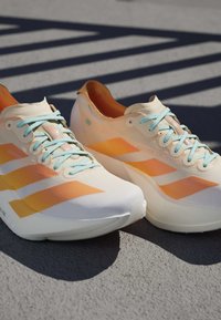 Een paar witte en oranje hardloopschoenen met lichtblauwe veters geplaatst op grijs trottoir met gestreepte schaduwen erboven.