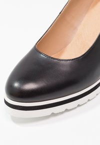 Chaussure en cuir noir à bout arrondi et design bas, avec une semelle blanche bordée d'une bande noire. Texture lisse.