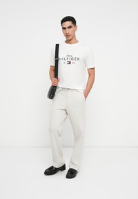 Bílý bavlněný tričko s námořnickou modrou a červenou grafikou, v kombinaci se světle béžovými kalhotami. Černá crossbody taška a černé mokasíny dotvářejí celý outfit.