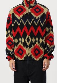 Pullover in fleece con collo alto, caratterizzato da un motivo geometrico in rosso, nero, beige e blu. Polsini elastici e chiusura con zip a metà. Texture morbida.