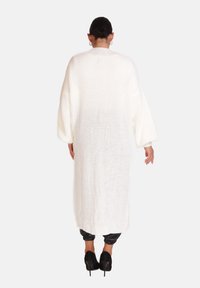 Langer, locker sitzender weißer Strickcardigan mit strukturiertem Muster, überschnittenen Schultern und weiten Ärmeln, kombiniert mit schwarzen schmal geschnittenen Hosen und hohen Schuhen.