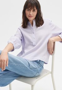 Camicia di cotone viola chiaro, con texture, colletto alla coreana, bottoni frontali e maniche arrotolate, abbinata a jeans azzurri, seduta su una sedia.