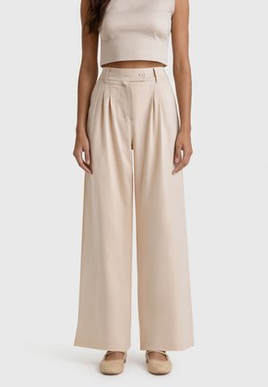 Donna che indossa pantaloni beige a vita alta, gamba larga con pieghe e top corto smanicato abbinato, completati da ballerine beige.