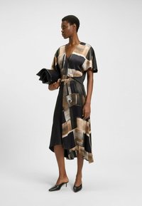 Robe asymétrique noire avec un motif marron et beige. Tissu léger avec un décolleté en V et des manches courtes. Le modèle tient une pochette noire.