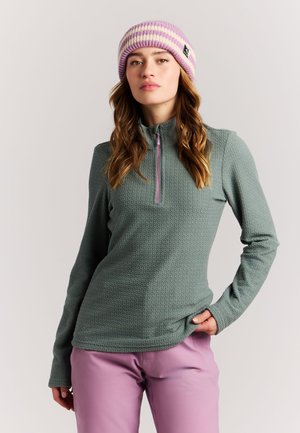 Vrouw draagt een groen textuur kwartritspullover, roze broek en een gestreepte roze-witte gebreide muts, staand met één hand in de zak.