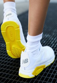 Baskets blanches avec des accents jaunes, semelle en caoutchouc texturée, tige en mesh et fonction "ABZORB". Portées avec des chaussettes blanches montantes.