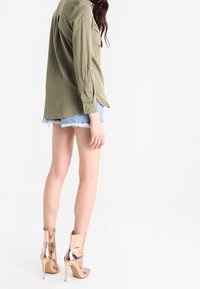 Donna che indossa una maglietta a maniche lunghe verde oliva, pantaloncini di jeans azzurri e stivaletti alla caviglia con tacco alto color oro rosa metallico lucido, in piedi su sfondo bianco.