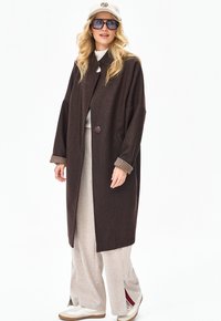 Blondynka w dużym ciemnobrązowym płaszczu, szerokich beżowych spodniach, białych trampkach, białej czapce w panterkę oraz dużych kwadratowych okularach przeciwsłonecznych.