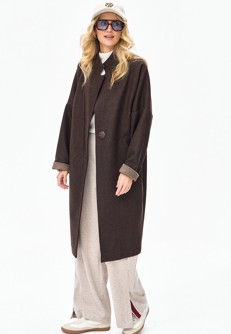 Blondynka w dużym ciemnobrązowym płaszczu, szerokich beżowych spodniach, białych trampkach, białej czapce w panterkę oraz dużych kwadratowych okularach przeciwsłonecznych.