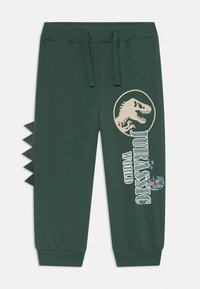 Pantaloni della tuta verdi con vita elastica, grafica di dinosauro e testo "Jurassic World" sul fronte, e spuntoni in tessuto lungo il lato.
