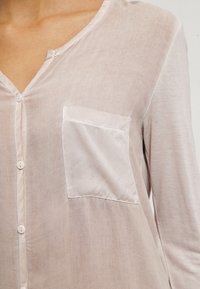 Blouse beige clair avec une texture douce et transparente. Elle comporte une fermeture boutonnée sur le devant, une petite poche poitrine et un col simple.
