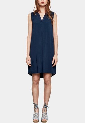 Robe sans manches bleu marine avec un décolleté en V, en tissu léger, longueur genou avec un ourlet arrière légèrement plus long, assortie à des sandales à talons bleu clair.